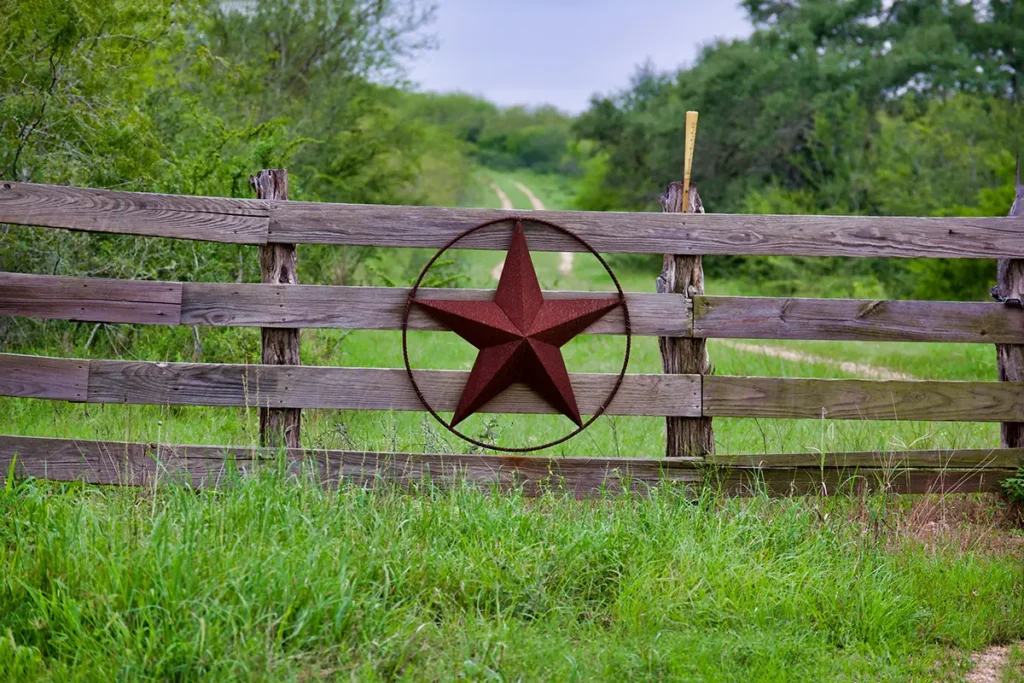 A metal Texas star hanging on a rustic wooden fence in a grassy area with trees in the background